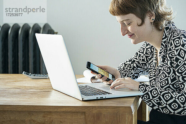 Lächelnde Frau mit Smartphone und Laptop bei der Arbeit von zu Hause aus