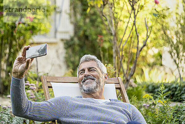 Lächelnder reifer Mann  der ein Selfie mit seinem Smartphone macht  während er auf einem Liegestuhl sitzt