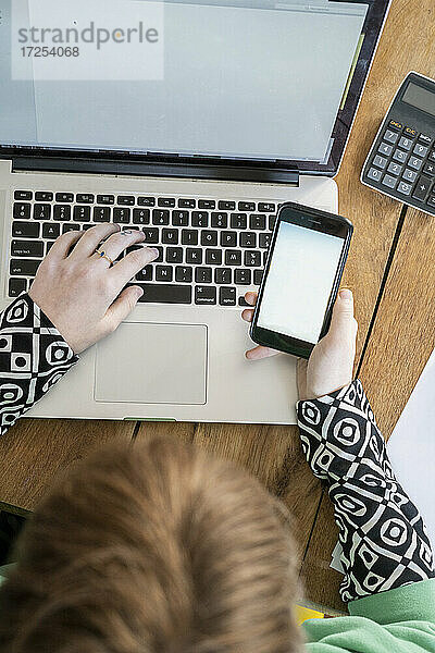 Draufsicht auf eine Frau  die ein Smartphone benutzt  während sie an einem Laptop arbeitet