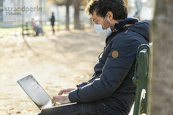 Seitenansicht eines Mannes mit Gesichtsschutzmaske  der in einem öffentlichen Park einen Laptop benutzt