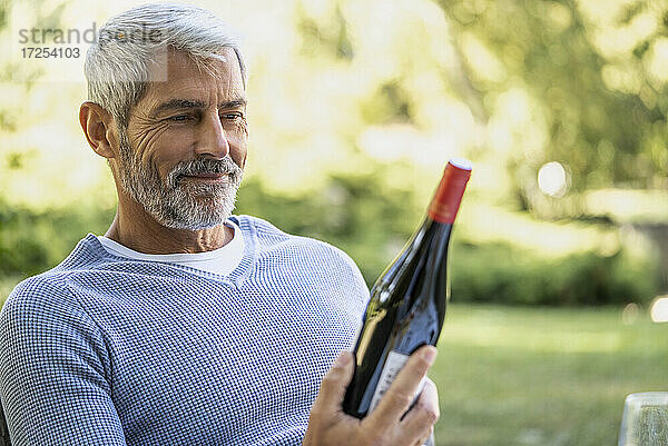 Lächelnder reifer Mann  der eine Weinflasche betrachtet  während er auf einem Stuhl sitzt