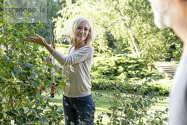 Lächelnde reife Frau mit Gartenschere sieht ihren Mann im Garten an