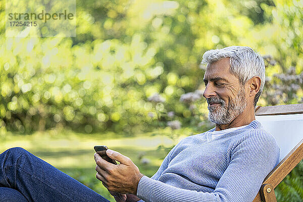Lächelnder reifer Mann  der ein Smartphone benutzt  während er auf einem Liegestuhl sitzt