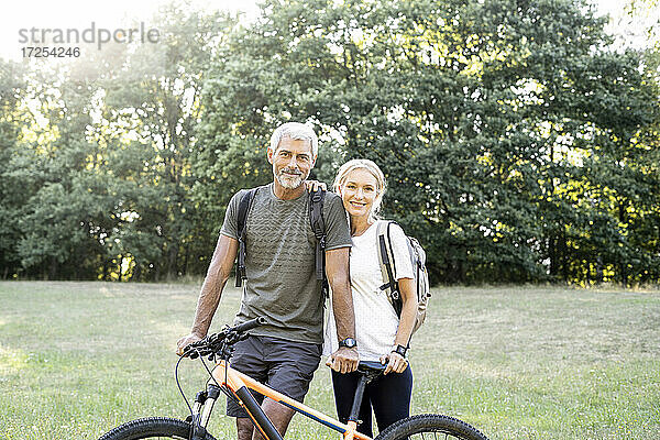 Porträt eines lächelnden reifen Paares mit Fahrrad im Wald stehend