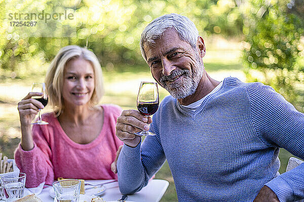 Porträt eines lächelnden reifen Paares mit Weingläsern in der Hand  das am Tisch sitzt