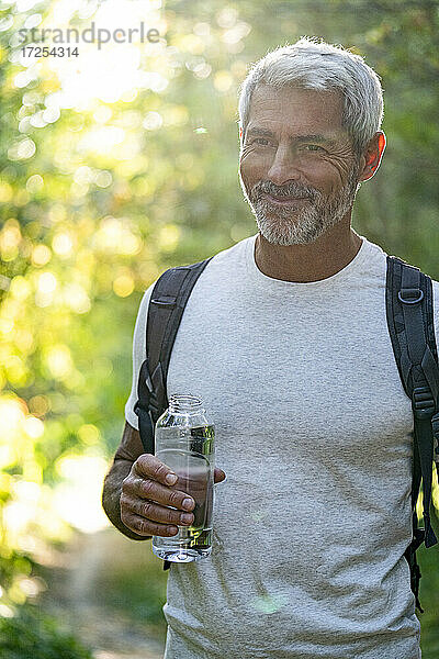 Porträt eines lächelnden reifen Mannes  der eine Wasserflasche im Wald hält