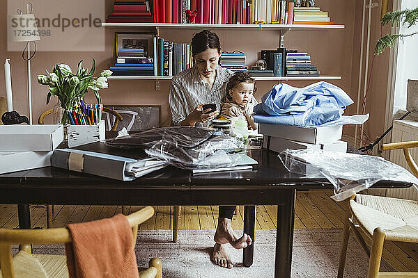 Arbeitende Mutter mit Smartphone beim Sitzen mit Baby Junge im Büro zu Hause