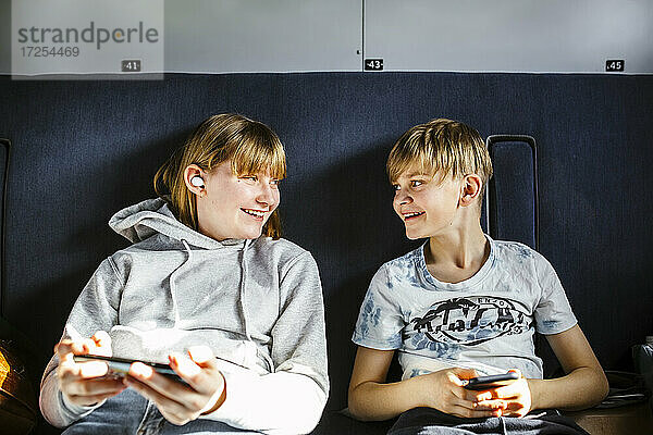 Lächelnde Bruder und Schwester mit Smartphones schauen sich an  während sie im Zug reisen
