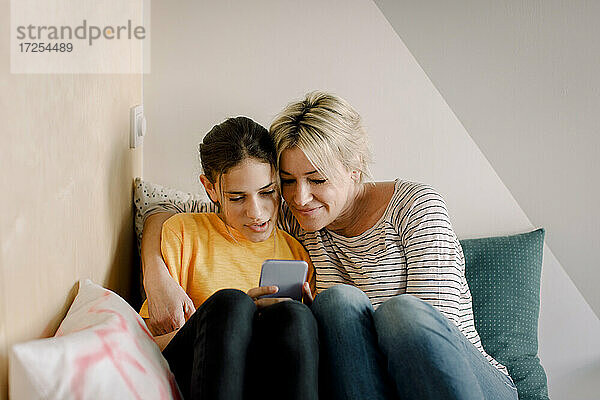 Reife Frau sitzt mit Tochter mit Smartphone im Schlafzimmer zu Hause