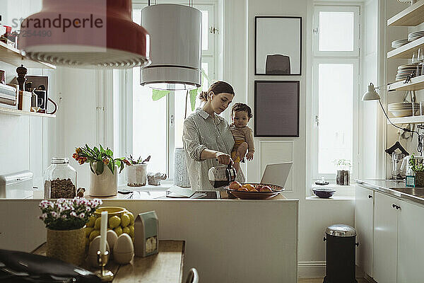 Weiblicher Profi gießt Kaffee ein  während er seinen kleinen Sohn in der Küche trägt