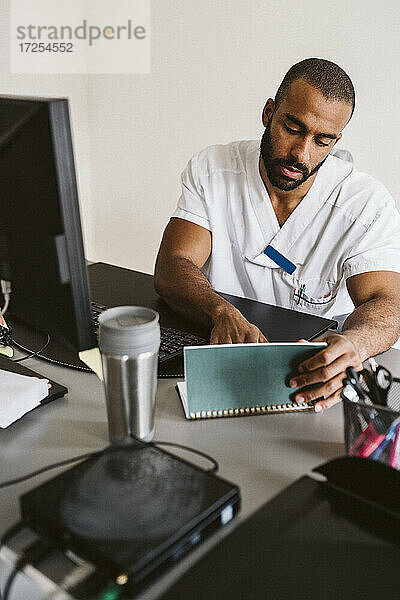 Männliche Gesundheitspflegekraft liest Krankenakte sitzend am Schreibtisch in medizinischer Klinik