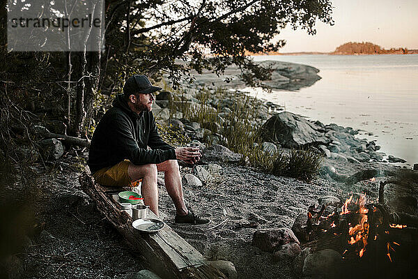 Mann in voller Länge  der wegschaut  während er auf einem Baumstamm am Lagerfeuer am Seeufer sitzt