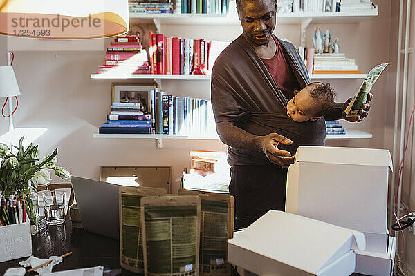 Männlicher Unternehmer  der Kisten untersucht  während er einen kleinen Jungen in der Trage im Büro zu Hause trägt