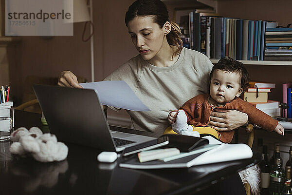 Weibliche Fachkraft liest Dokument  während sie mit männlichem Kleinkind zu Hause sitzt
