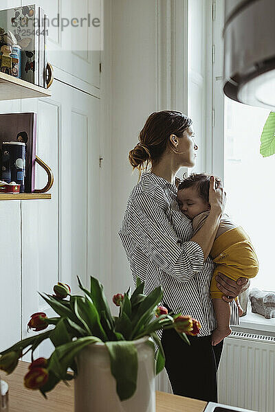 Mutter schaut durch das Fenster  während sie ein männliches Kleinkind zu Hause trägt