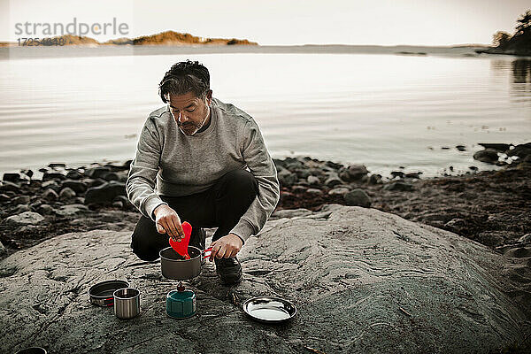 Reifer Mann in voller Länge beim Kochen von Essen auf einem Campingkocher am Seeufer