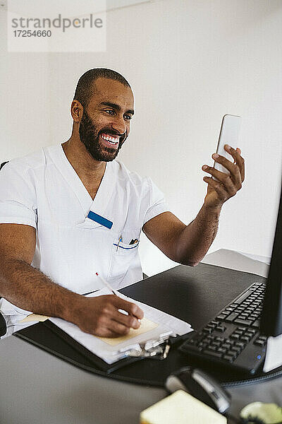 Fröhliche männliche Arzt Beratung durch Smartphone beim Schreiben am Schreibtisch