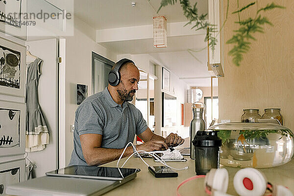 Geschäftsmann mit Kopfhörern  der an einem Laptop arbeitet  während er an einer Kücheninsel sitzt