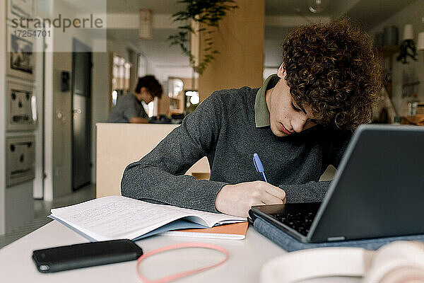 Teenager-Junge mit braunen lockigen Haaren macht Hausaufgaben beim Sitzen am Tisch