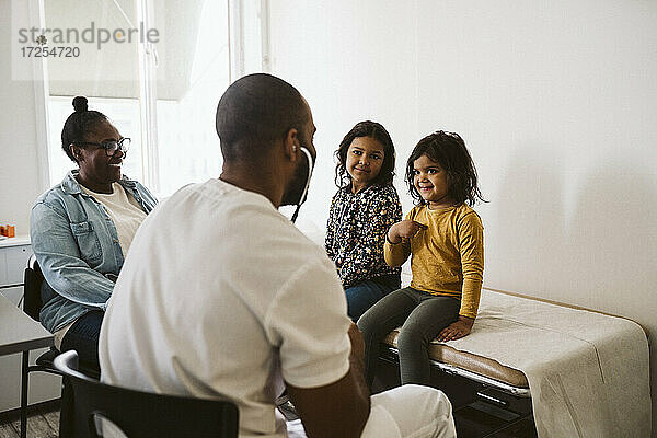 Lächelndes Mädchen  das bei seiner Schwester sitzt und gestikuliert  während es einen männlichen Arzt in einer medizinischen Klinik ansieht