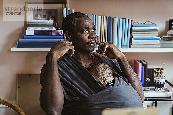 Vater nachdenklich beim Sitzen mit Sohn in Babytrage zu Hause