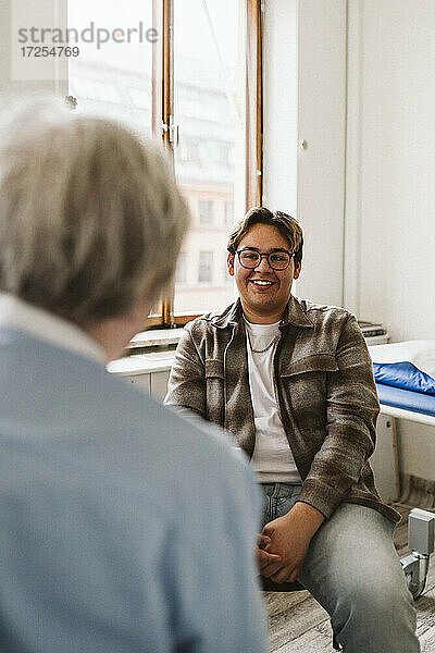 Lächelnder männlicher Patient  der einen älteren weiblichen medizinischen Experten während einer Konsultation in der Klinik ansieht