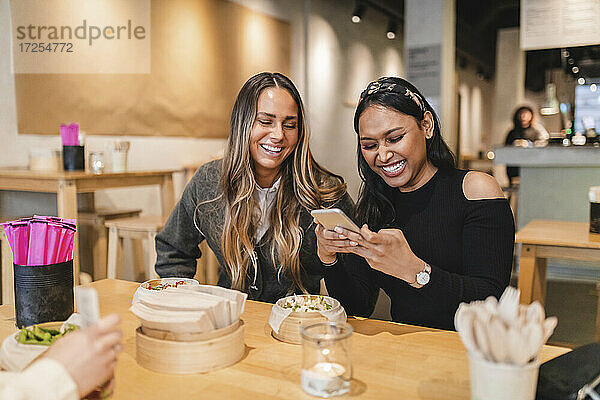 Lächelnde Freundinnen  die ein Smartphone benutzen  während sie im Restaurant sitzen