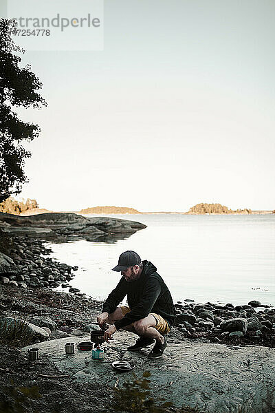 Mann in voller Länge beim Kochen von Essen  während er auf einem Felsen am Seeufer hockt