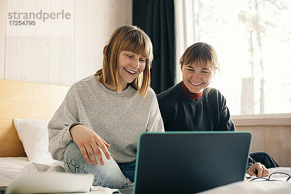 Lächelnde Mutter und Tochter sitzen mit Laptop während Homeschooling im Schlafzimmer