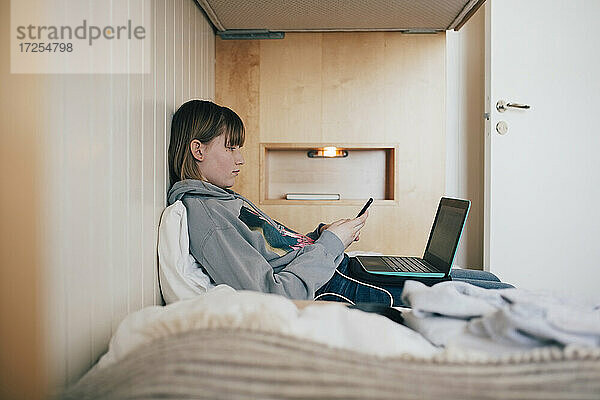 Teenager-Mädchen sitzt mit Laptop mit Smartphone in Ferienhaus