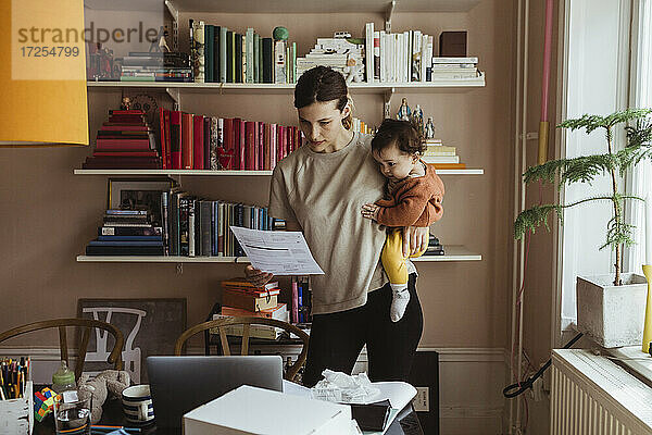 Weiblicher Profi  der ein Dokument liest  während er seinen Sohn im Büro zu Hause trägt