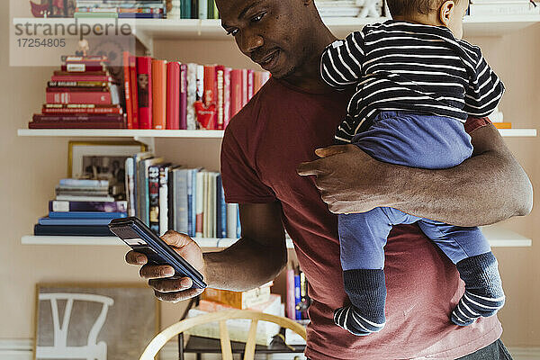 Vater verwendet Smartphone  während er einen kleinen Jungen zu Hause trägt