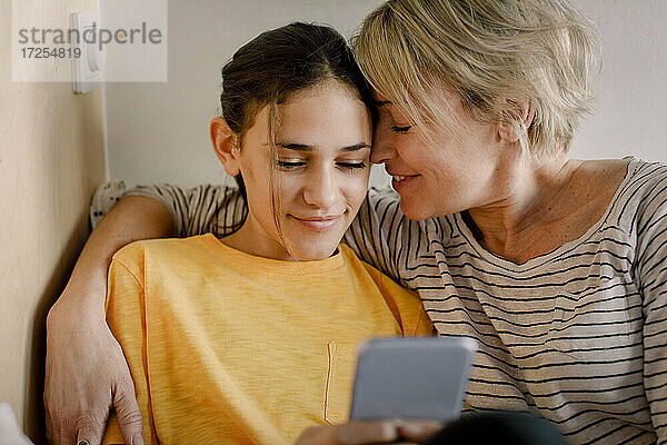 Glückliche Mutter umarmt Tochter mit Mobiltelefon im Schlafzimmer
