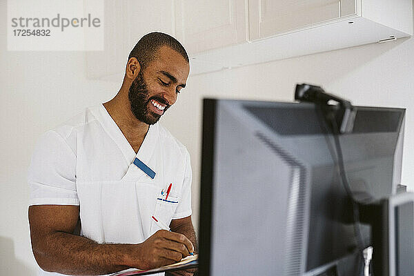 Lächelnde männliche Pflegekraft beim Schreiben  während sie den Patienten per Videoanruf berät