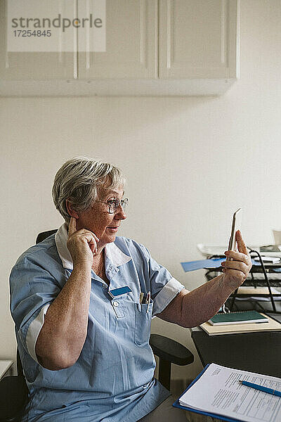 Älterer weiblicher Arzt  der die Ohren berührt  während er einen Patienten über ein Smartphone in einer medizinischen Klinik berät