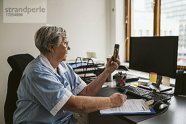 Älterer medizinischer Experte berät Patienten per Videoanruf  während er am Schreibtisch ein Rezept schreibt