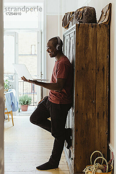 Lächelnder männlicher Unternehmer  der einen Laptop benutzt  während er sich zu Hause an einen Schrank lehnt