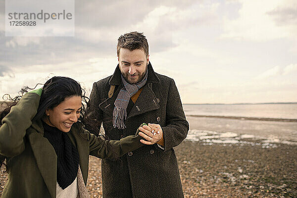 Glückliches Paar in Wintermänteln  die Hände haltend  am Strand spazierend