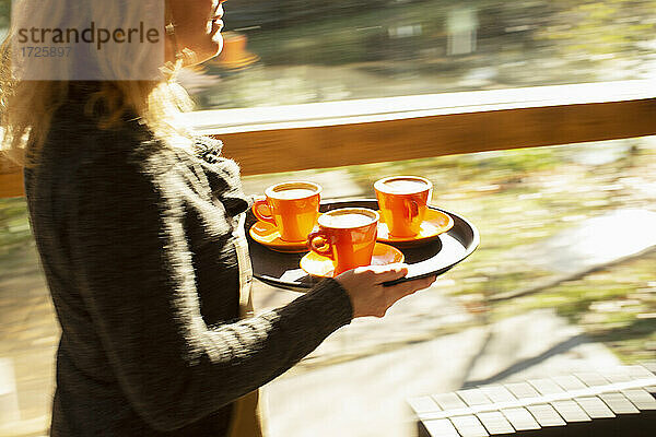 Weibliche Barista trägt Tablett von Kaffeetassen entlang sonnigen Café Fenster