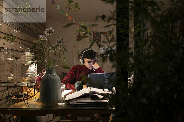 Konzentrierter junger Mann mit Kopfhörern arbeitet spät am Laptop im Heimbüro