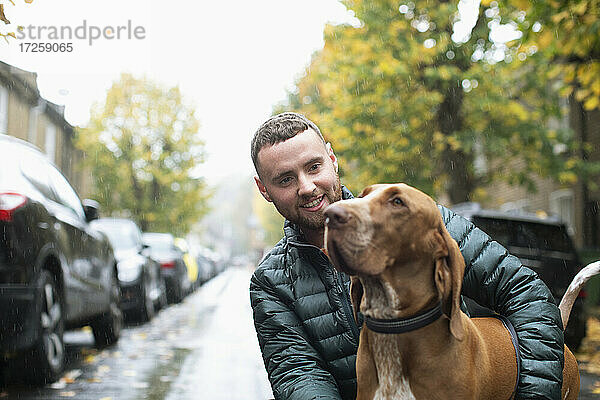 Glücklicher junger Mann mit Hund auf nassen städtischen Straße