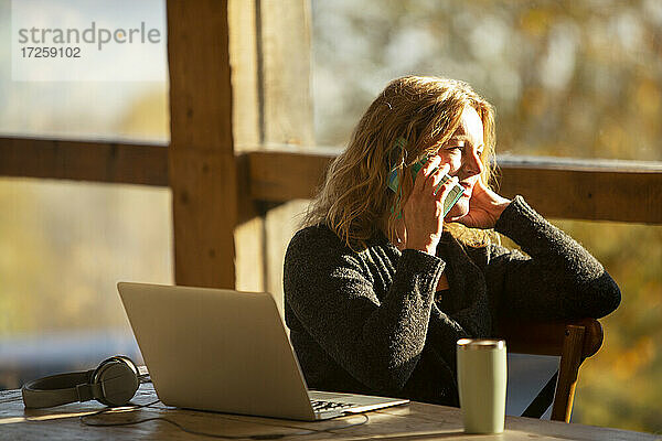 Geschäftsfrau im Gespräch auf Smartphone am Laptop in sonnigen Cafe