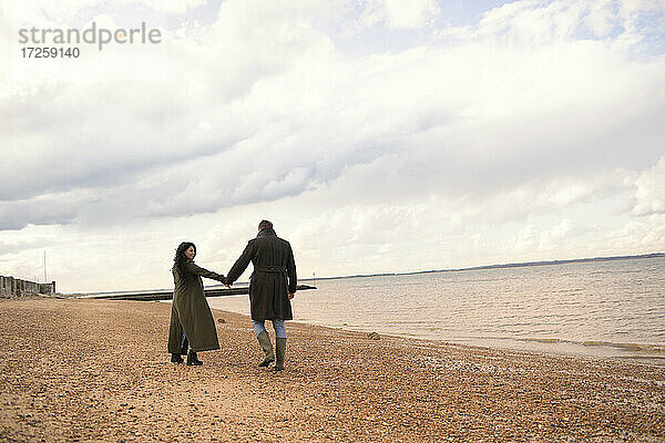 Paar in Wintermänteln  die sich an den Händen halten und am Strand spazieren gehen