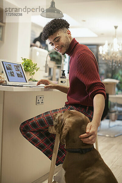 Junger Mann arbeitet von zu Hause am Laptop und streichelt Hund in der Küche