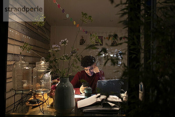 Junger Mann mit Smartphone arbeitet nachts von zu Hause am Laptop