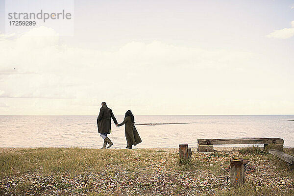 Paar in Wintermänteln  die sich an den Händen halten und am Strand spazieren gehen