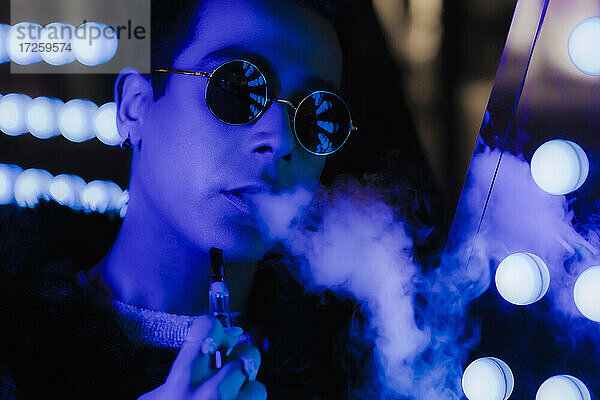 Portrait kühlen jungen Mann in Sonnenbrille vaping in dunklen Neon. Nachtclub