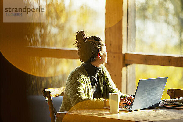 Frau arbeitet am Laptop und schaut aus dem Fenster in einem sonnigen Cafe