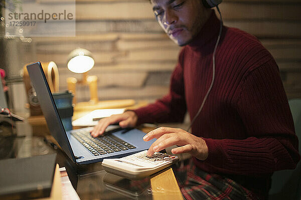 Junger Mann mit Kopfhörer mit Taschenrechner am Laptop im Home-Office
