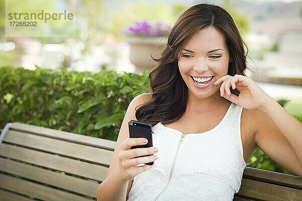 Attraktive lächelnde junge erwachsene Frau schreibt auf Handy im Freien auf einer Bank